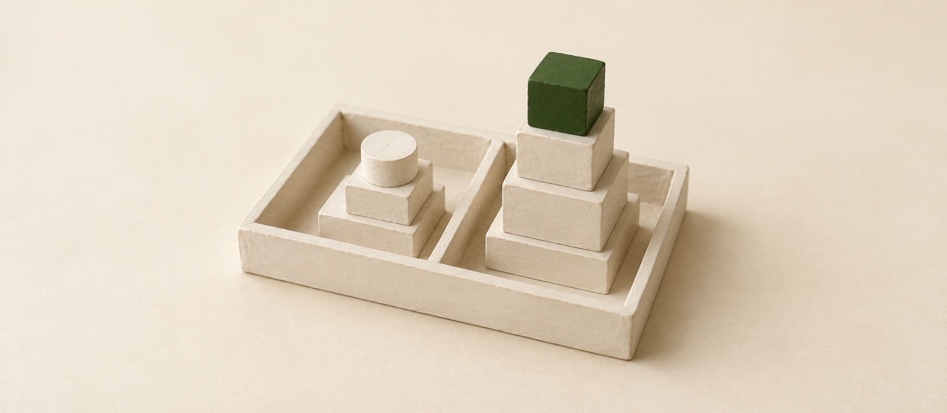 A cream study model of a shallow tray divided in two, a short stack of plain blocks on one side and a taller stack on the other, with the top block of the taller stack in green.