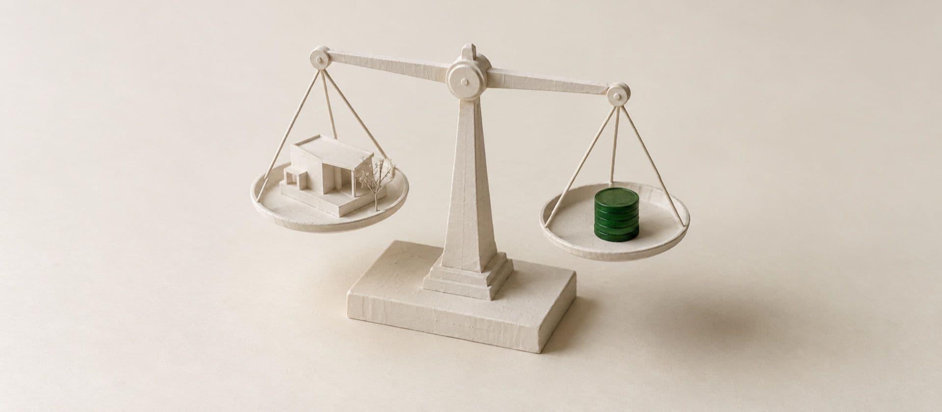 A cream study model of a two-pan balance scale with a tiny shop model on one pan and a short stack of coins on the other, with the stack of coins in green.