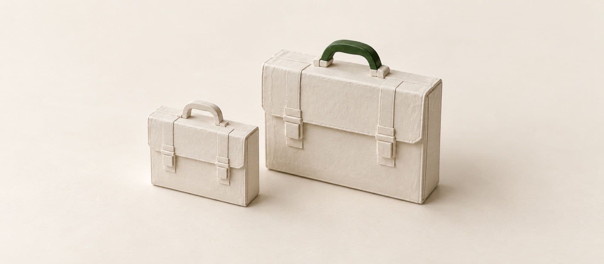 A cream study model of two closed briefcases standing side by side, one small and one large, with the handle of the larger briefcase in green.