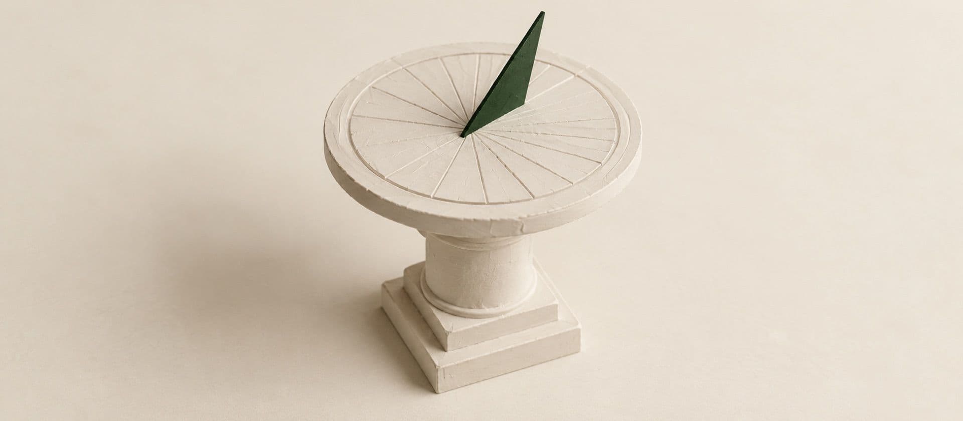 A cream study model of a garden sundial on a short pedestal, with the sundial's pointer in green.