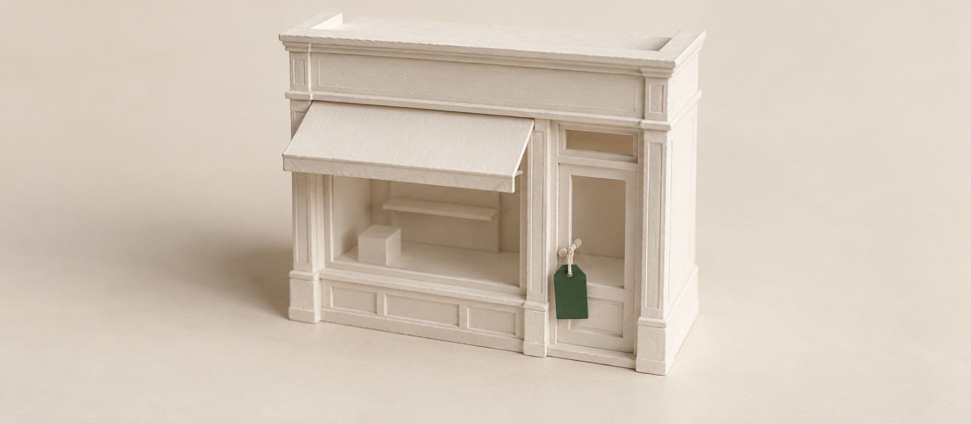 A cream study model of a small model of a shopfront with a blank luggage-style price tag tied to its door handle by a string, with the price tag in green.