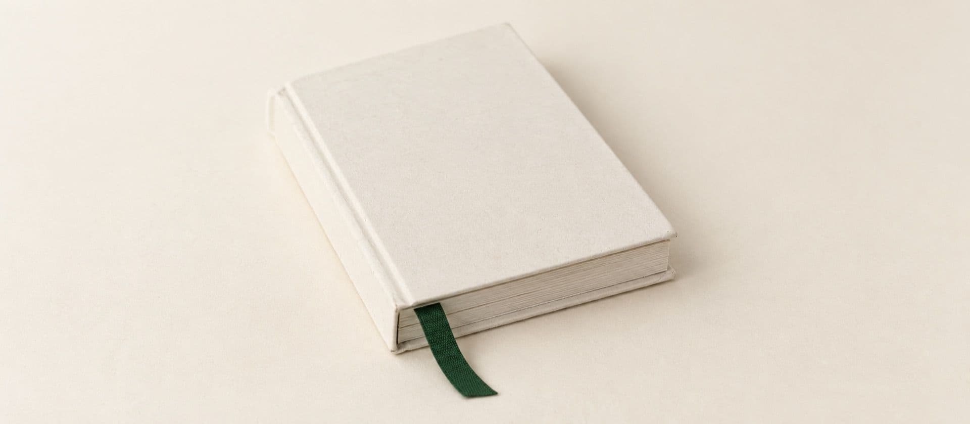 A cream study model of a closed bound booklet with a blank cover and a ribbon bookmark hanging from its pages, with the ribbon in green.
