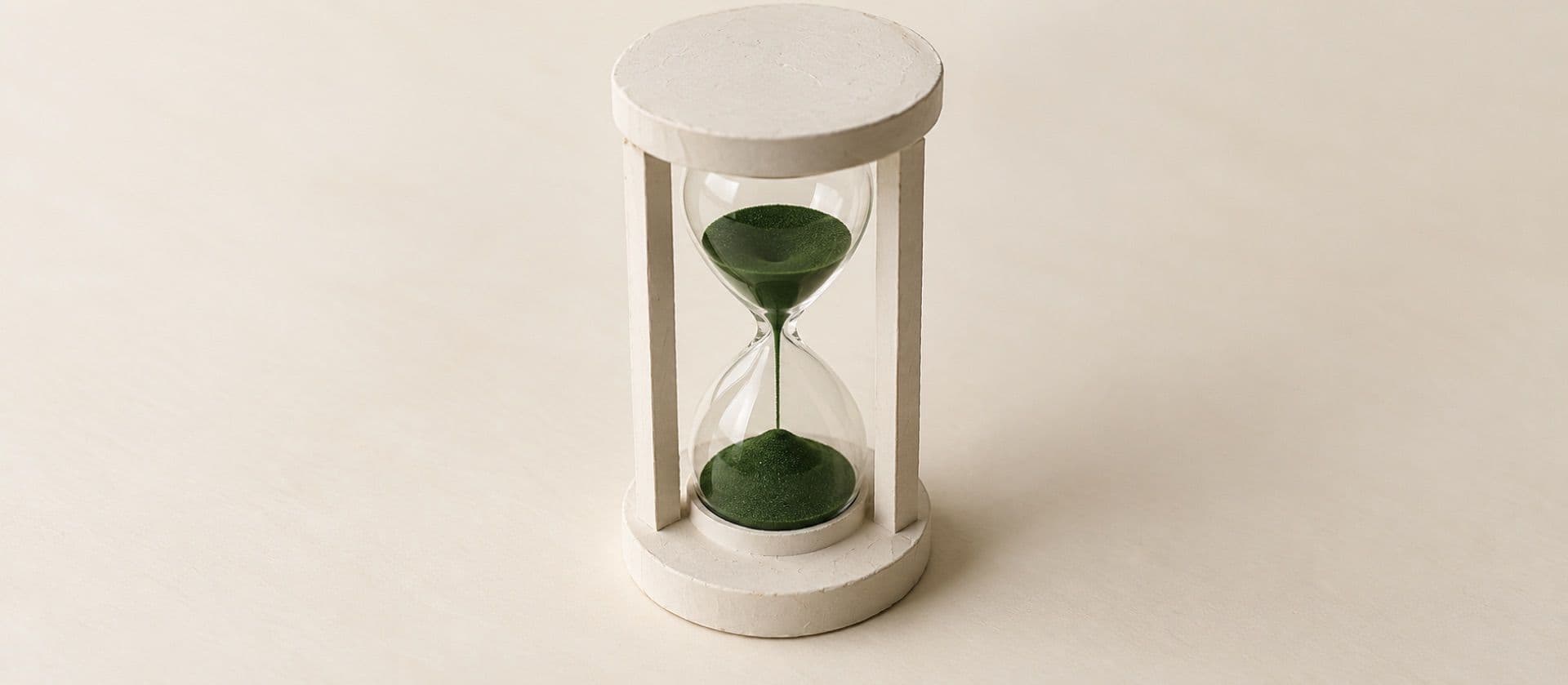 A cream study model of an hourglass standing upright with sand in both bulbs, with the sand in green.