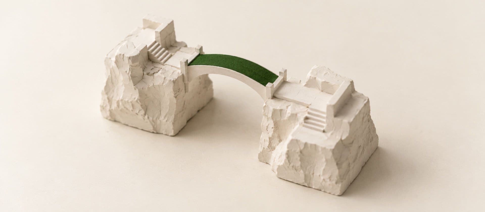 A cream study model of two separate stone blocks joined by a small arched footbridge between them, with the bridge deck in green.